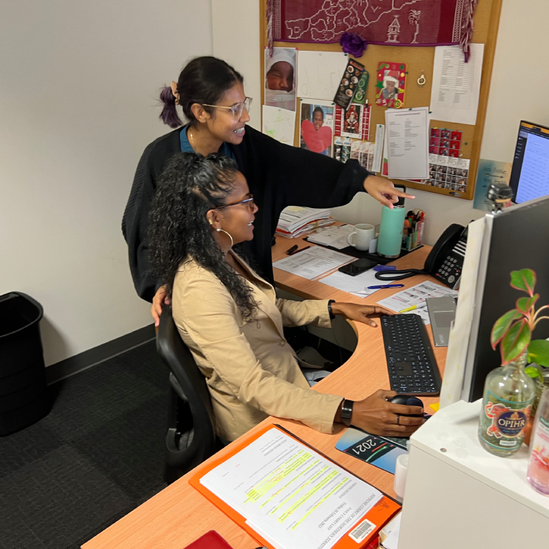 Two women behind an office desk, looking at a computer monitor and working on a task together.