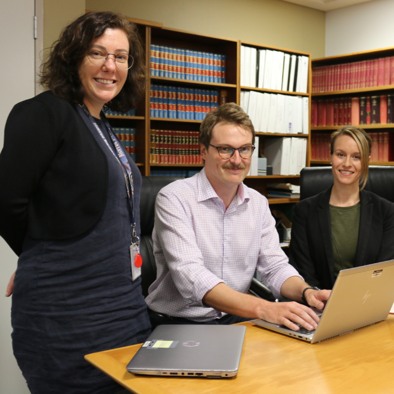 A woman standing on the left, a man sitting in the middle with their hands on a laptop, and a woman sitting on the right smiling, all in front of bookshelves filled with law books.